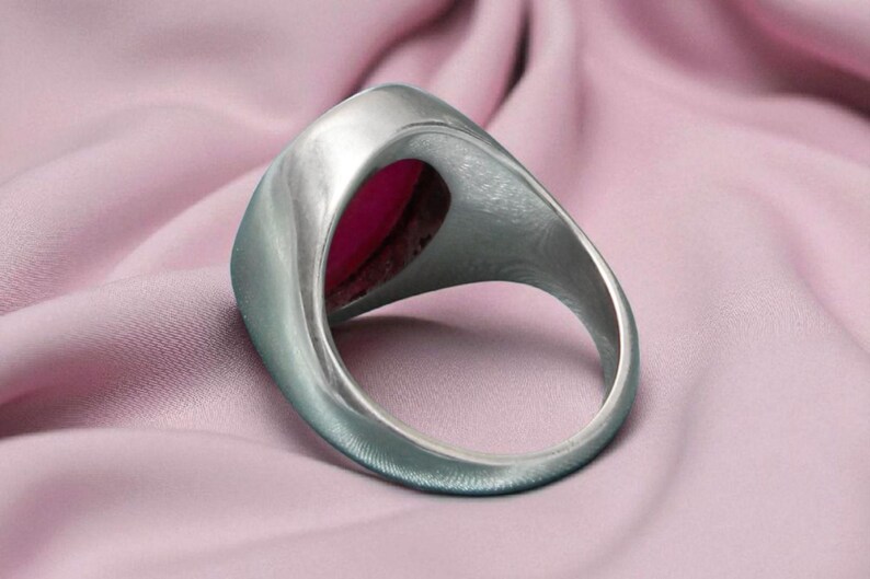 May include: A silver ring featuring a large, oval, magenta gemstone. The ring is set against a soft, pink fabric, accentuating the jewellery's design and colour. The ring's band is thick and smooth.