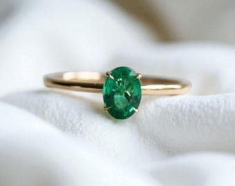 May include: A gold ring featuring a vibrant green oval-cut gemstone. The ring's band is a polished gold, and the gemstone is set with delicate prongs. The ring is displayed on a white, textured surface, highlighting its elegant design.