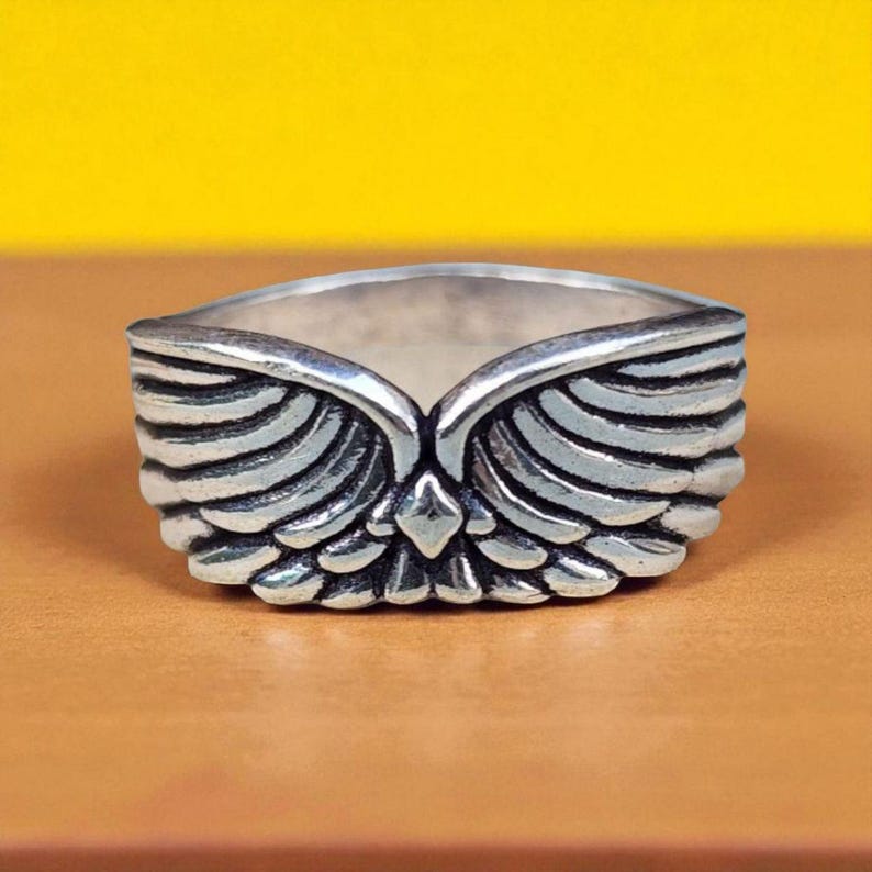 Op de afbeelding: Een zilverkleurige ring met een gedetailleerd vleugelontwerp. De ring heeft een centraal punt en gelaagde veerd details, waardoor een driedimensionaal effect ontstaat. De ring staat tegen een gele en bruine achtergrond.
