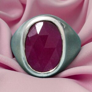 May include: A silver ring featuring a large, faceted, oval ruby gemstone. The ring is set against a soft pink fabric background, creating a contrast between the cool metal and the warm gemstone.