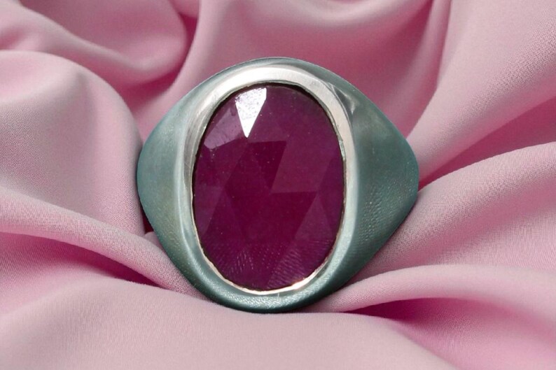 May include: A silver ring featuring a large, faceted, oval ruby gemstone. The ring is set against a soft pink fabric background, creating a contrast between the cool metal and the warm gemstone.