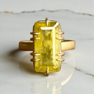 Könnte beinhalten: Ein goldfarbener Ring mit einem großen, rechteckigen gelben Edelstein. Der Edelstein ist in einem goldenen Band mit dekorativen Elementen gefasst. Der Ring wird auf einer Marmoroberfläche präsentiert. Der Text "Zivira Jewels" ist auf dem Ring sichtbar.