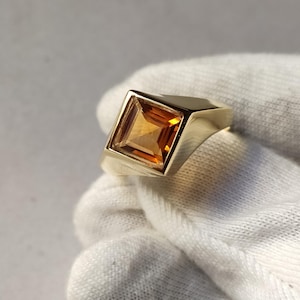 May include: A gold ring with a square, amber-coloured gemstone. The gemstone is set in a square bezel, and the ring has a simple, polished band. The ring is shown against a white background.