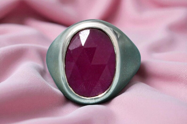 May include: A silver ring featuring a large, oval-cut, deep red gemstone. The ring has a wide band and a bezel setting. The background is a soft, pink fabric, creating a luxurious feel.