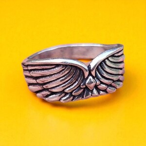 Op de afbeelding: Een zilverkleurige ring met een gedetailleerd vleugelontwerp. De ring heeft twee vleugels die in het midden samenkomen, met een klein diamantvormig accent. De ring staat tegen een heldergele achtergrond.