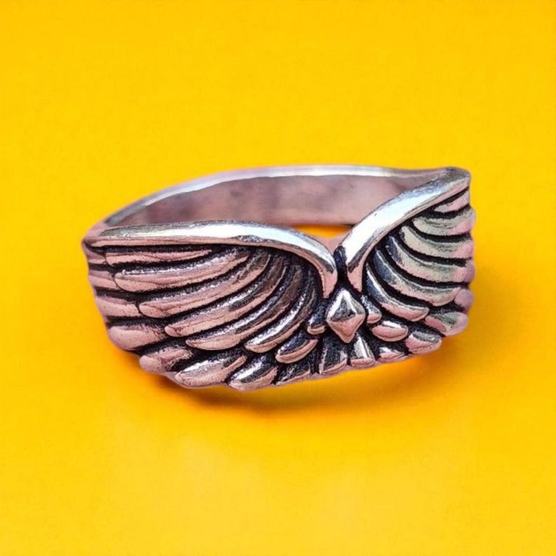 Op de afbeelding: Een zilverkleurige ring met een gedetailleerd vleugelontwerp. De ring heeft twee vleugels die in het midden samenkomen, met een klein diamantvormig accent. De ring staat tegen een heldergele achtergrond.