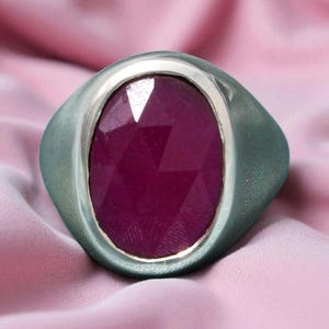 May include: A silver ring featuring a large, oval-cut, deep red gemstone. The ring has a wide band and a bezel setting. The background is a soft, pink fabric, creating a luxurious feel.