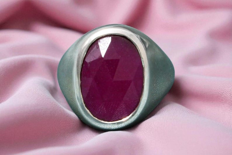 May include: A silver ring featuring a large, oval-cut, deep red gemstone. The ring has a wide band and a bezel setting. The background is a soft, pink fabric, creating a luxurious feel.