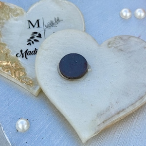 May include: Two heart-shaped magnets. One is white with gold leaf accents and the name "Madi" printed on it. The other is white with a black magnet attached. Two faux pearls are visible.