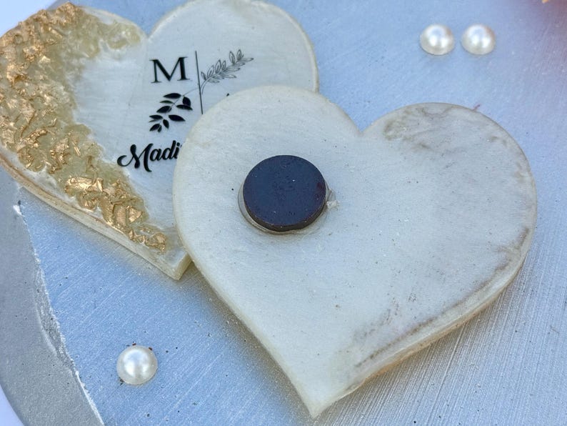 May include: Two heart-shaped magnets. One is white with gold leaf accents and the name "Madi" printed on it. The other is white with a black magnet attached. Two faux pearls are visible.