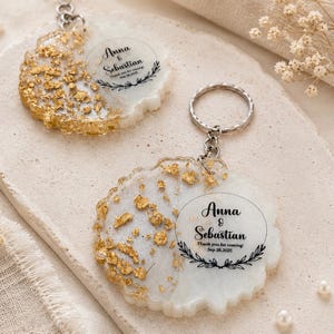May include: Two round resin keychains featuring a white base with gold leaf flakes on one side. Each keychain has a silver metal ring and is printed with the text Anna & Sebastian, Thank you for coming, Sep 28, 2025, and a leaf wreath.