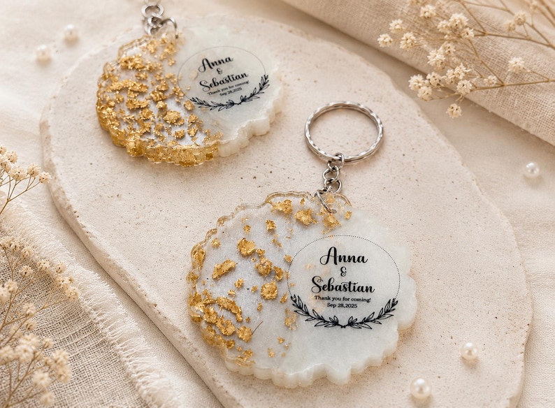 May include: Two round resin keychains featuring a white base with gold leaf flakes on one side. Each keychain has a silver metal ring and is printed with the text Anna & Sebastian, Thank you for coming, Sep 28, 2025, and a leaf wreath.