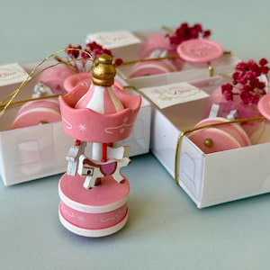 May include: A pink and white carousel ornament with a gold top, featuring a small horse figure. The ornament is displayed in front of two white gift boxes, each tied with gold string and decorated with pink wax seals reading Thank You.