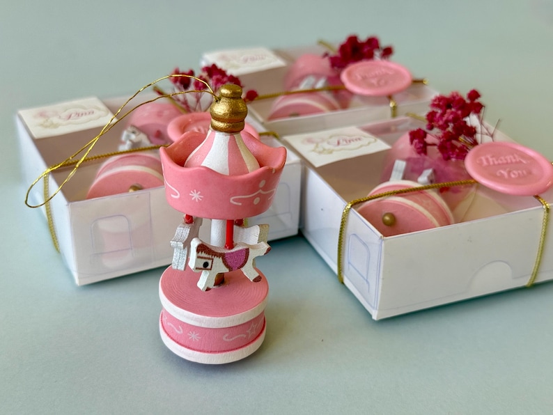 May include: A pink and white carousel ornament with a gold top, featuring a small horse figure. The ornament is displayed in front of two white gift boxes, each tied with gold string and decorated with pink wax seals reading Thank You.
