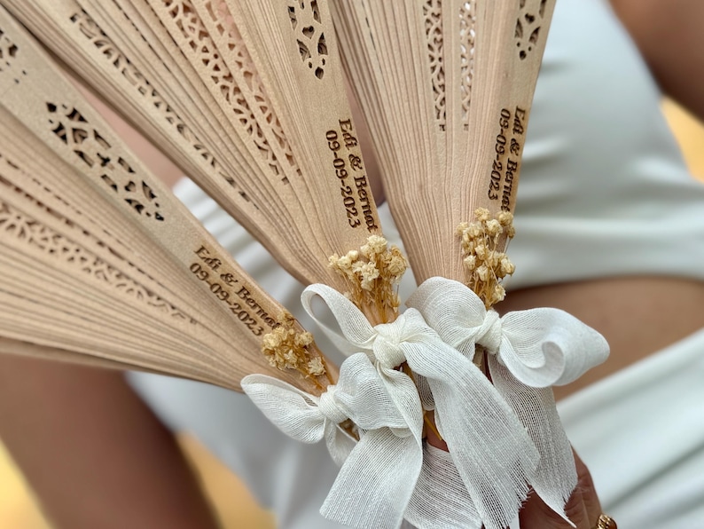 May include: Close-up of several wooden fans with intricate cut-out designs. Each fan is tied with a white ribbon and adorned with dried flowers. The fans are personalized with the text "Edi & Bernat 09-09-2023".
