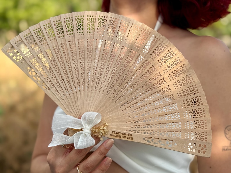 May include: A light brown wooden hand fan with intricate cut-out designs. The fan is tied with a white ribbon and small dried flowers. Text on the fan reads "Ed & Bernat 09-09-2023".