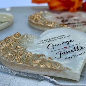 May include: Heart-shaped wedding favors with gold accents and personalized text. The favors are white with gold leafing on the edges and feature the names "George" and "Janette" with the date "May 27, 2024".