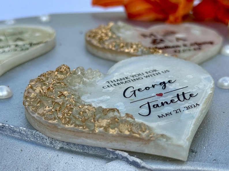 May include: Heart-shaped wedding favors with gold accents and personalized text. The favors are white with gold leafing on the edges and feature the names "George" and "Janette" with the date "May 27, 2024".