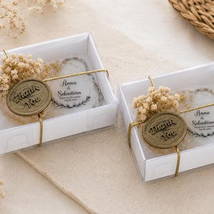 May include: Two clear square gift boxes containing round white keychains with the text Anna & Sebastian and Sep 08, 2025. Each box is tied with gold string and features a round bronze-coloured wax seal with the text Thank You and dried flowers.