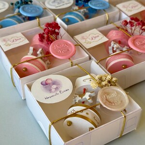 May include: A collection of white gift boxes containing miniature carousel music boxes with horses. Each box is tied with gold string, a wax seal reading Thank You, a small dried flower sprig, and a personalized circular tag with names or graphics.