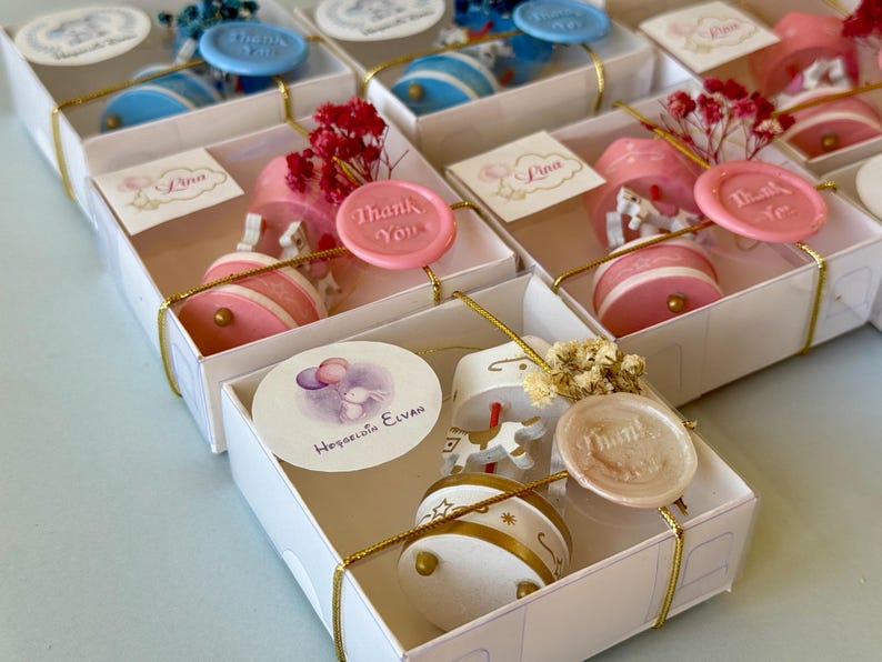 May include: A collection of white gift boxes containing miniature carousel music boxes with horses. Each box is tied with gold string, a wax seal reading Thank You, a small dried flower sprig, and a personalized circular tag with names or graphics.