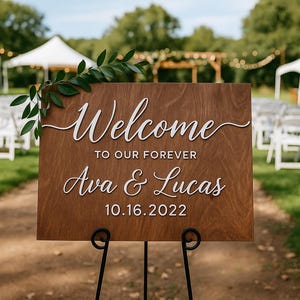 Puede incluir: Un letrero de bienvenida de boda de madera con letras blancas que dice "Welcome to our forever Ava & Lucas 10.16.2022". El letrero se muestra en un soporte de metal negro, con vegetaci&oacute;n en la parte superior izquierda. El fondo muestra un lugar de boda.