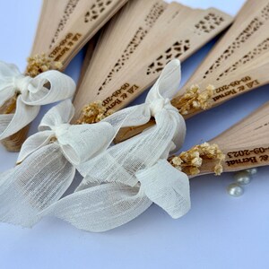 May include: Wooden fans with decorative cutouts, tied with ivory ribbons and dried floral accents. Each fan is engraved with a date and names. A few pearls are scattered near the fans. Ideal for wedding favors.
