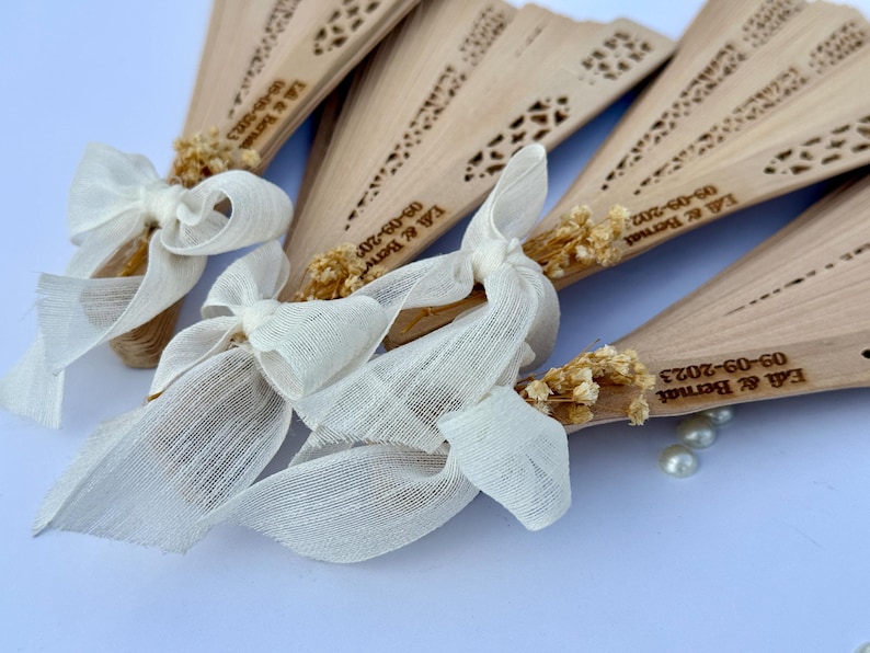 May include: Wooden fans with decorative cutouts, tied with ivory ribbons and dried floral accents. Each fan is engraved with a date and names. A few pearls are scattered near the fans. Ideal for wedding favors.