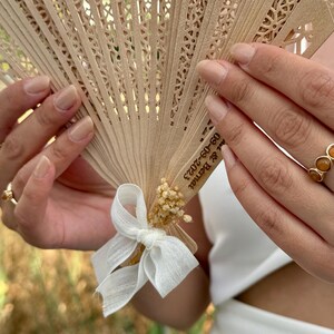 May include: A beige wooden fan with intricate cut-out designs, held open. The fan is adorned with a white ribbon and small dried flowers. The text "09-09-2023 & Bernat" is printed on the fan. A gold ring with three orange stones is visible.