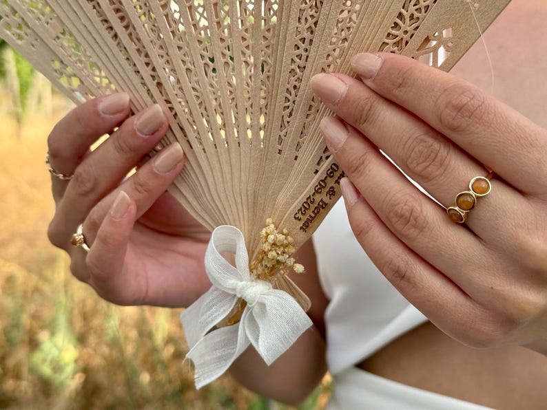 May include: A beige wooden fan with intricate cut-out designs, held open. The fan is adorned with a white ribbon and small dried flowers. The text "09-09-2023 & Bernat" is printed on the fan. A gold ring with three orange stones is visible.