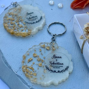 May include: Two circular resin keychains with uneven, wavy edges featuring embedded gold leaf flakes. Each keychain displays the printed text Anna & Sebastian, Thank you for coming! Sep 28, 2025, framed by a delicate black laurel leaf graphic.