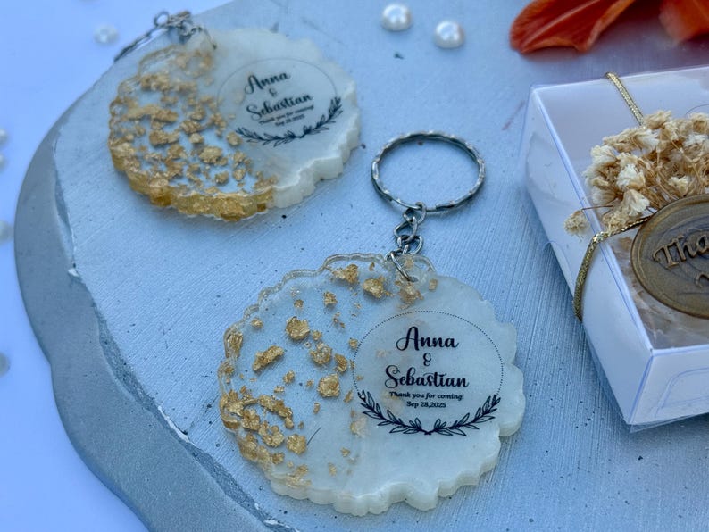 May include: Two circular resin keychains with uneven, wavy edges featuring embedded gold leaf flakes. Each keychain displays the printed text Anna & Sebastian, Thank you for coming! Sep 28, 2025, framed by a delicate black laurel leaf graphic.