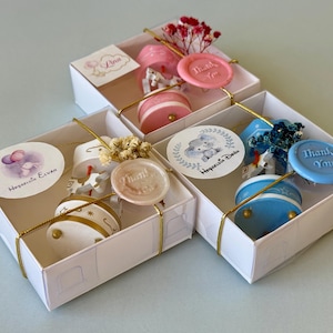 May include: Three white square gift boxes containing small carousel-style ornaments in pink, white, and blue. Each box is tied with gold string, a wax seal embossed with "Thank You," and a round sticker featuring illustrations and text.