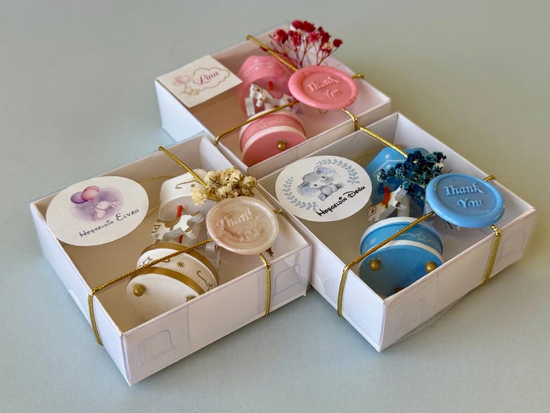 May include: Three white square gift boxes containing small carousel-style ornaments in pink, white, and blue. Each box is tied with gold string, a wax seal embossed with "Thank You," and a round sticker featuring illustrations and text.
