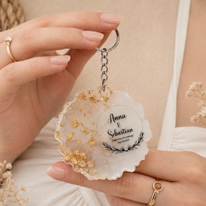 May include: A round resin keyring with a wavy edge, featuring a white base with gold leaf flakes on one side. The keyring includes a silver metal ring and chain, with black text reading Anna & Sebastian, Thank you for coming!, Sep 28, 2025.