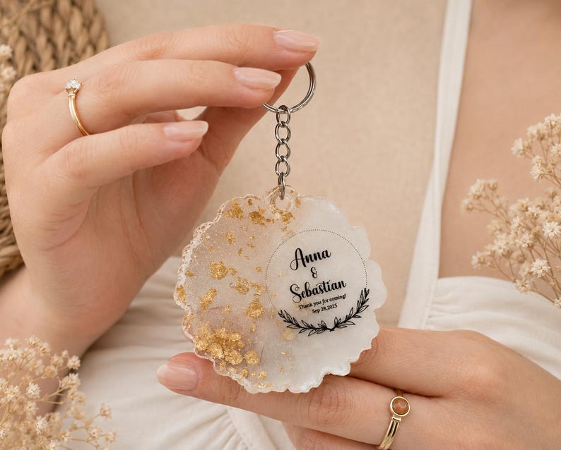 May include: A round resin keyring with a wavy edge, featuring a white base with gold leaf flakes on one side. The keyring includes a silver metal ring and chain, with black text reading Anna & Sebastian, Thank you for coming!, Sep 28, 2025.