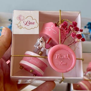 May include: A small pink carousel music box inside a clear square gift box. The carousel features a white horse with pink accents. A pink wax seal with the text Thank You is attached to the front with gold string, alongside dried pink flowers.