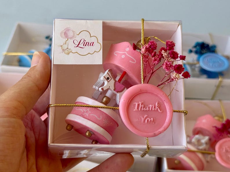 May include: A small pink carousel music box inside a clear square gift box. The carousel features a white horse with pink accents. A pink wax seal with the text Thank You is attached to the front with gold string, alongside dried pink flowers.