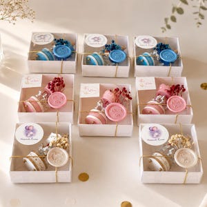May include: A collection of seven square white gift boxes, each containing a small glass bottle with dried flowers and a wax seal stamped with the words Thank You. The boxes are arranged in three rows, featuring blue, pink, and white color themes.