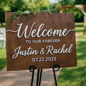 Puede incluir: Un letrero de boda de madera con letras blancas que dice "Welcome to our forever Justin & Rachel 07.22.2023". El letrero est&aacute; sobre un soporte de metal negro, con vegetaci&oacute;n en la parte superior. El fondo muestra una zona de c&eacute;sped y sillas blancas.