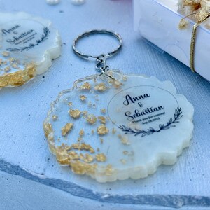 May include: A round resin keyring with a scalloped edge, featuring embedded gold leaf flakes and a printed design that reads Anna & Sebastian, Thank you for coming, Sep 28, 2025, framed by a black botanical wreath graphic and a metal key ring.