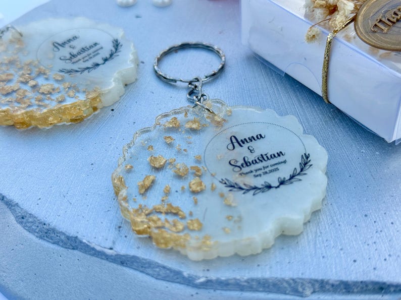 May include: A round resin keyring with a scalloped edge, featuring embedded gold leaf flakes and a printed design that reads Anna & Sebastian, Thank you for coming, Sep 28, 2025, framed by a black botanical wreath graphic and a metal key ring.