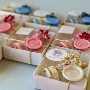 May include: A collection of small white gift boxes, each tied with gold string and decorated with dried flowers and a wax seal. Some seals are pink, blue, or white, featuring the text Thank You. A round sticker with balloons and the text Hosgeldin Elvan is visible.