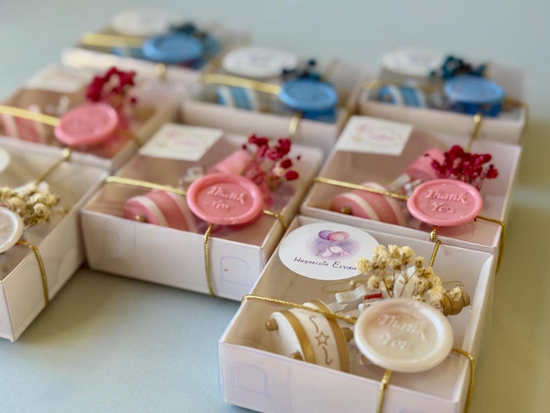 May include: A collection of small white gift boxes, each tied with gold string and decorated with dried flowers and a wax seal. Some seals are pink, blue, or white, featuring the text Thank You. A round sticker with balloons and the text Hosgeldin Elvan is visible.