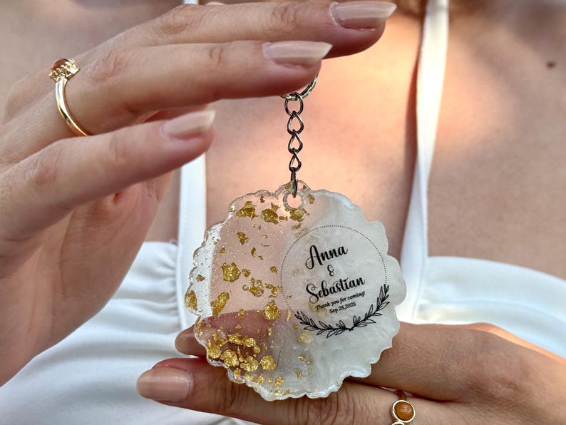 May include: A circular resin keyring with a wavy edge, featuring a white and translucent pink marbled design with embedded gold leaf flakes. The keyring includes black printed text that reads Anna & Sebastian, Thank you for coming!, Sep 28, 2025, and a decorative leaf motif.