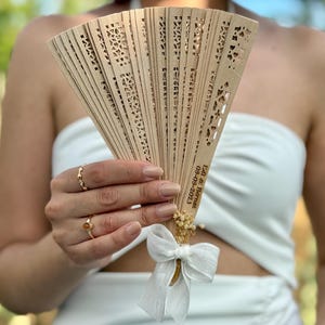 May include: A light brown wooden fan with intricate cut-out designs. The fan is adorned with a white ribbon, dried flowers, and text that reads "Edi & Barnet 09-09-2023". The fan is held in front of a person wearing a white strapless top.