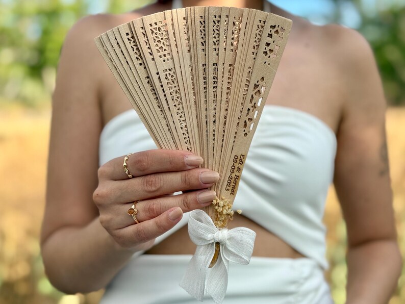 May include: A light brown wooden fan with intricate cut-out designs. The fan is adorned with a white ribbon, dried flowers, and text that reads "Edi & Barnet 09-09-2023". The fan is held in front of a person wearing a white strapless top.