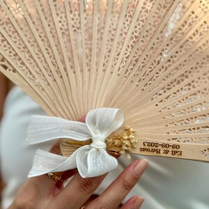 May include: A light brown wooden fan with intricate cut-out designs. The fan is adorned with a white ribbon bow and small dried flowers. The text "Edi & Bernat 09-09-2023" is visible on the fan.