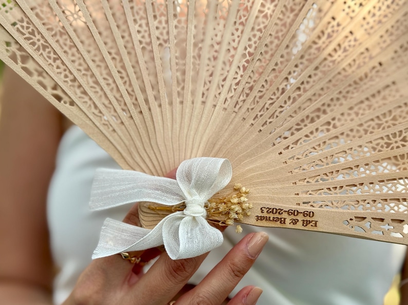 May include: A light brown wooden fan with intricate cut-out designs. The fan is adorned with a white ribbon bow and small dried flowers. The text "Edi & Bernat 09-09-2023" is visible on the fan.