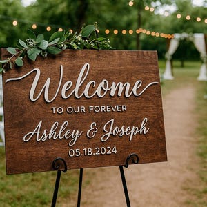 Puede incluir: Un letrero de boda de madera con las palabras "Welcome to our Forever Ashley & Joseph 05.18.2024" en escritura blanca. El letrero est&aacute; decorado con vegetaci&oacute;n y se encuentra sobre un caballete de metal negro en un entorno exterior cubierto de hierba.